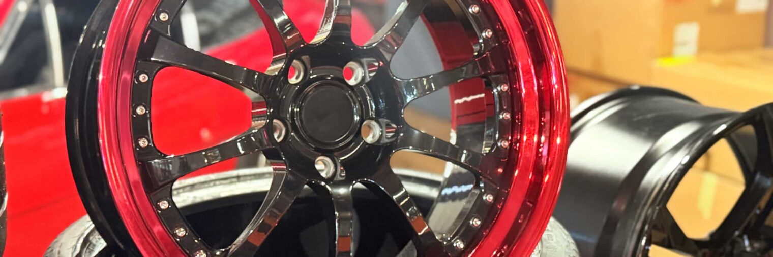 A close-up of a black and red custom car wheel rim resting on a tire, highlighting quality craftsmanship in the automotive aftermarket sector, with another rim and cardboard boxes visible in the background.