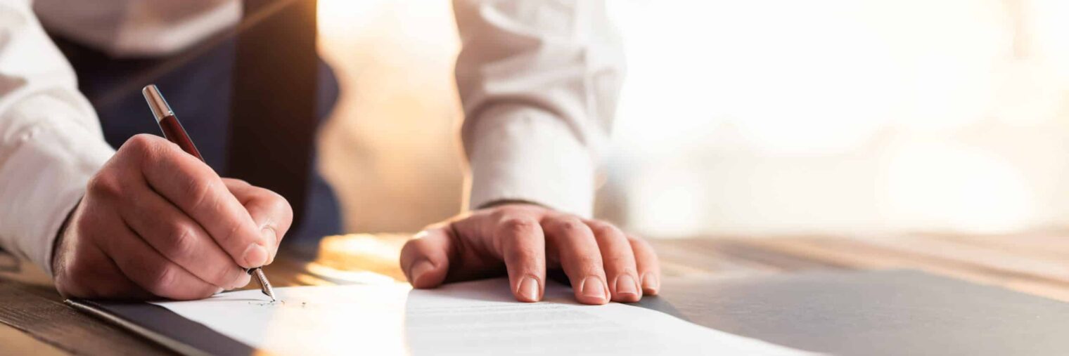 A person in business attire signs a document on a desk, holding a pen in one hand and resting the other on the paper, symbolizing key moments in sell side advisory or Mergers & Acquisitions Advisory Services within Investment Banking.