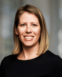 A woman with straight, shoulder-length blonde hair wearing a black top, smiling at the camera against a blurred background.