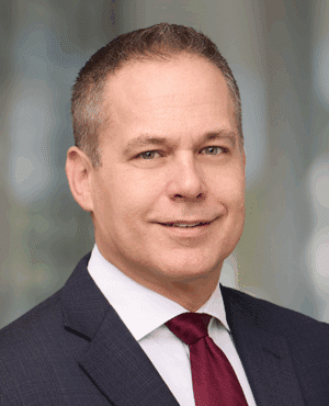 A man in a dark suit, white shirt, and red tie, with short brown hair, posing for a professional headshot against a blurred background.
