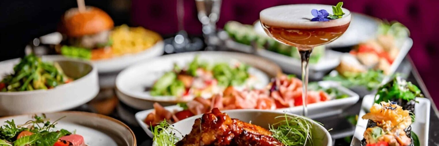 A table set with various plated dishes—including meat, salad, sushi, and cocktails—stands out against a purple upholstered background, capturing the lively spirit of the restaurant market.