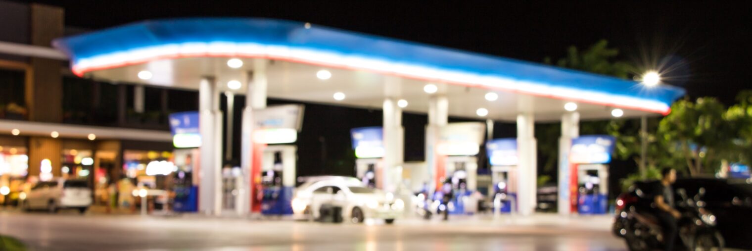 A blurred image of a gas station at night, with cars parked at fuel pumps and bright lights illuminating the canopy, reflecting the modern look often seen after convenience store acquisitions.