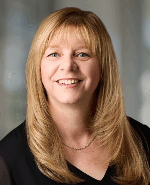 A woman with straight blonde hair and bangs, wearing a black top and a thin necklace, smiles at the camera against a blurred indoor background.