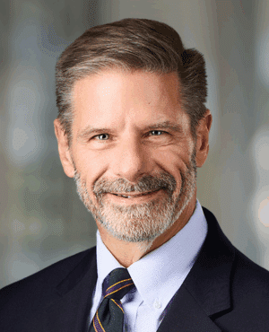 Middle-aged man with short gray hair and a beard, wearing a dark suit, white shirt, and striped tie, smiling at the camera with a blurred background.